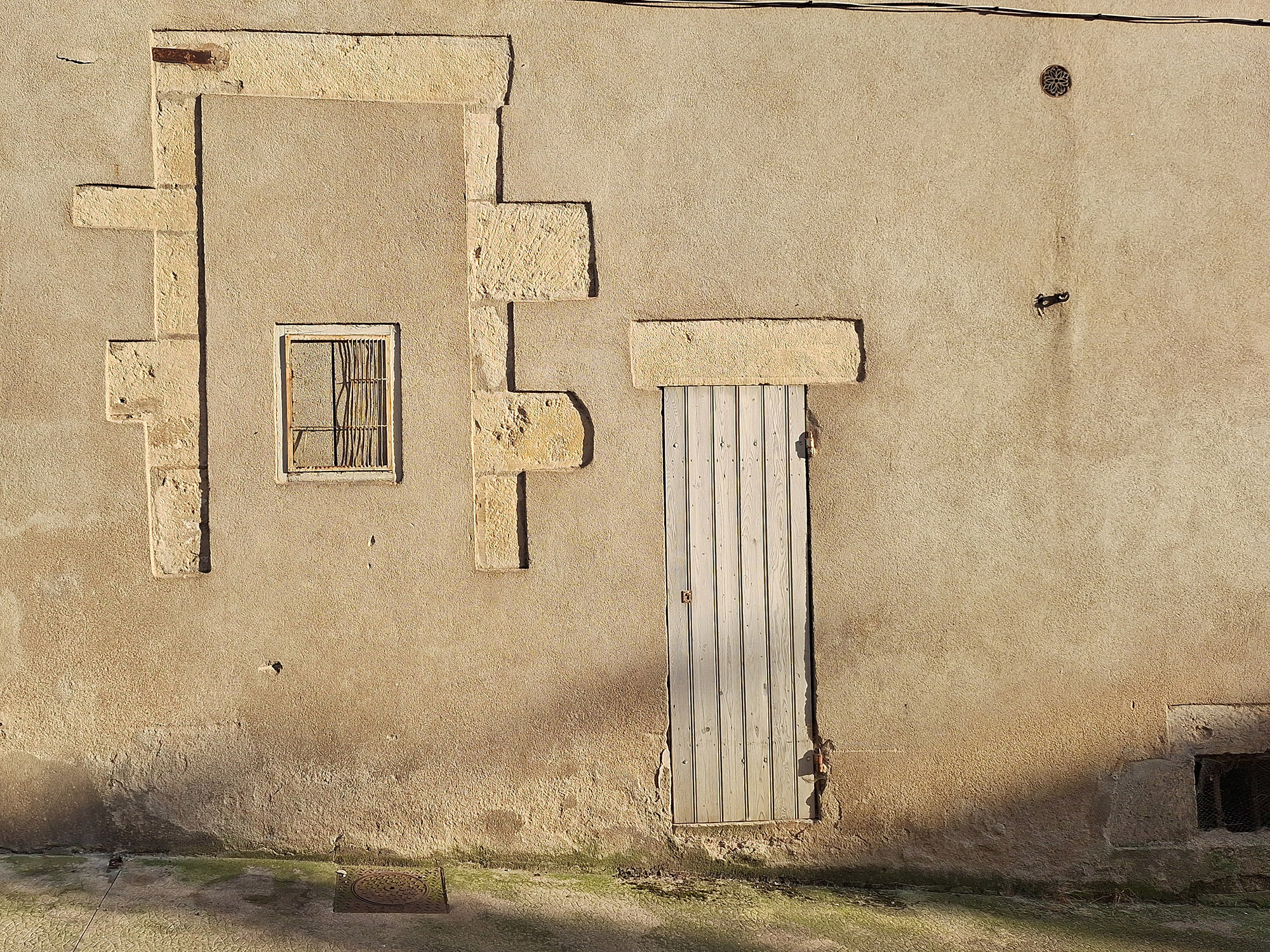 Weathered wall with a sealed former window protected by bent metal bars beside a narrow pale wooden door, photographed in low evening sunlight in Avallon.