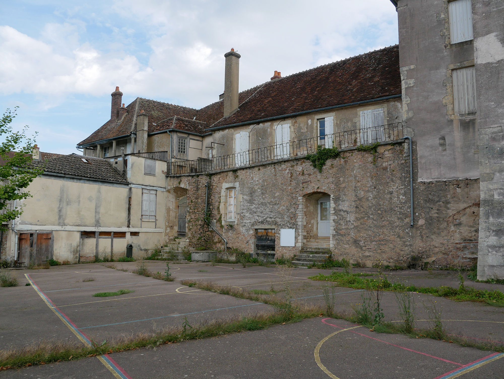 Ancienne cour d’école avec marquages sportifs effacés et végétation traversant le sol, au pied de bâtiments fermés à Avallon.