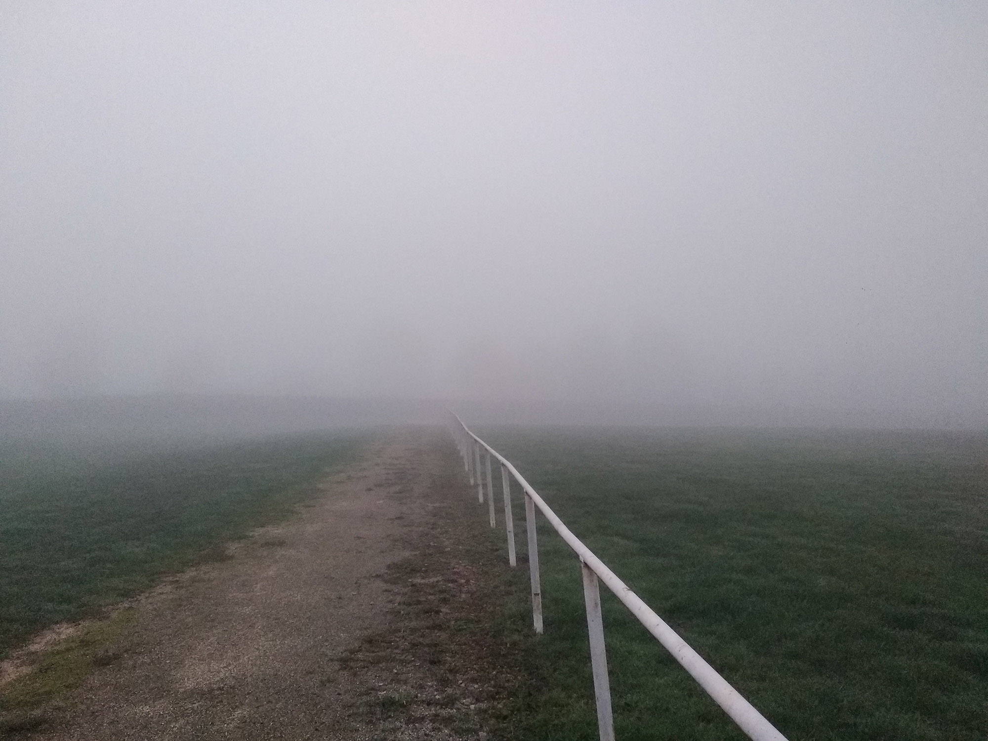 A road disappearing into dense fog beyond visibility, bordered by a white railing across an open field near Avallon.