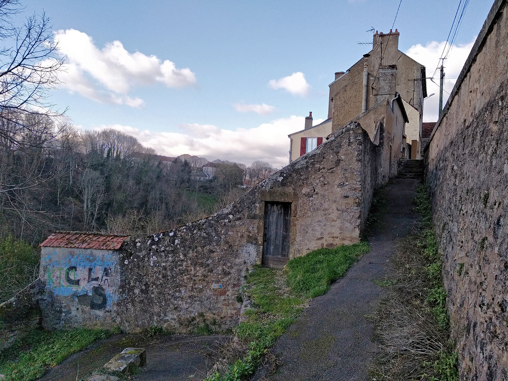 Narrow stone pathway descending between old walls and houses in Avallon, opening toward a wooded valley under a cloudy sky.